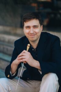 Portrait of conductor Christian Olson smiling while sitting on outside steps holding a baton